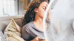 bir elinde telefon bir eliyle yüzünü kapatmış ve gözleri kapalı bir uzun kıvırcık saçlı bir kadın koltukta yaslanmış ve fan önünde serinlemeye çalışıyor