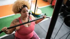 A foto de Strock mostra uma pessoa com uma tatuagem de flor no ombro direito vestindo roupas esportivas vermelhas e rosa puxando para baixo uma barra de pesos lateral em uma academia.