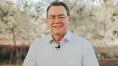 Um homem de meia-idade, Augusto Cury aparece sentado ao ar livre, diante de um fundo com árvores e vegetação desfocados, vestindo uma camisa social clara e óculos de armação fina, com um pequeno microfone preso à altura do peito. Ele sorri para a câmera.