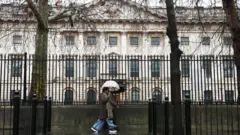 白い大きな建物の前の黒い柵に沿って、傘を差した人が歩いている