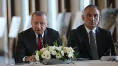Cumhurbaşkanı Recep Tayyip Erdoğan ve Turizm Bakanı Mehmet Nuri Ersoy bir masada oturuyor, önlerinde çiçek var.