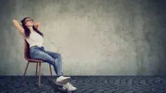 Young woman, wearing casual clothes, resting on a chair and holding her hands behind head - she looks like she is day-dreaming.