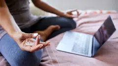 Closeup of a woman sitting cross-legged on her bed at home and using a streaming class on a laptop to help her meditate