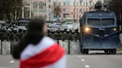 A protester with a flag stands in front of Belarusian policemen and a water cannon that block a road during a rally against government and President Lukashenko in Minsk, Belarus, 25 October 2020