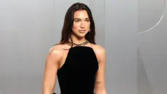 Dua Lipa pewnie pozuje przed kamerą, ubrana w czarną sukienkę z gorsetem i złoty, gruby naszyjnik. Jej długie, ciemne włosy są rozpuszczone.