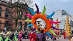 Di image show a vibrant parade wit various participants in colourful costumes. Di central focus na large, inflatable structure wey resemble a sun wit multicoloured rays, mounted on a purple platform. A performer stand inside di structure, dressed in a green and gold outfit. Surrounding di structure na participants in red outfits pushing di platform, and odas dey in green and purple costumes holding decorative staffs. Di parade dey take place on a street lined wit spectators, wit a historic brick building and a modern building in di background. Di scene dey lively and festive, indicating a celebration or festival.