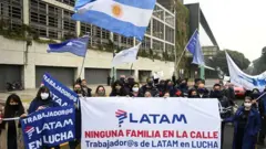 Dezenas de pessoas com máscaras contra o coronavírus, bandeiras e cartazes na rua, um delas com símbolo da Latam