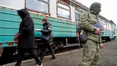 Homem fardado faz guarda em frente a trem, com duas mulheres caminhando atrás