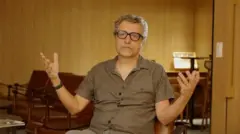 Kléber Mendonça Filho sentado, com os braços abertos