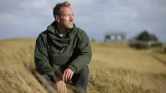 Ben Fogl u žitu, u zelenoj jakni i farmerkama, gleda u daljinu