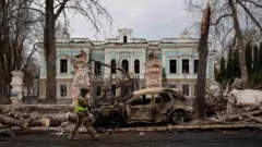 Сумы. На заднем плане старое здание с выбитыми стеклами, на переднем сгоревшая машина и обломки деревьев, мимо проходит человек в военной форме.
