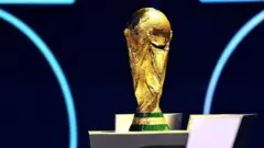サッカー男子W杯のトロフィー。台座は三角錐の下半分の形で、緑と金色に塗り分けられ、金色部分に「FIFA WORLD CUP」の文字が見える。トロフィー部分は金色で、4人の人物が大きなサッカーボールを掲げているような彫刻になっている