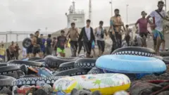 Boias salva-vidas de borracha estão dispostas em uma praia em frente a uma fila de migrantes que chegam a Ceuta.