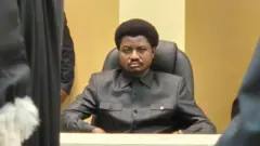 Constant Mutamba yambaye ishati y'umukara ikoze nk'ikoti, yicaye yegamye mu ntebe y'imisego y'umukara. Iruhande rwe haboneka igice cy'ikanzu y'umukara n'umweru yambawe n'umunyamategeko. Ifoto yo mu bubiko yakuwe kuri konti ya Constant Mutamba yo ku rubuga nkoranyambaga rwa X. 