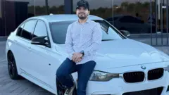César Gastélum sentado sobre un BMW blanco. Lleva una gorra negra