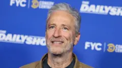 Jon Stewart, de jaqueta marrom e camiseta preta, com fundo de cor azul promocional do seu programa 'The Daily Show'
