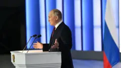 Putin gesticulando enquanto discursa em salão, com bandeira ao lado