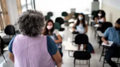 Professora em pé e de costas para a camera, olhando para alunos sentados em carteiras