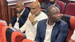 Nnamdi Kanu for court, e wear white Fendi designers sweater, and e sit in between DSS officers.