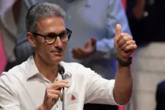 Zema sorrindo, segurando microfone com uma mãe e fazendo o sinal de positivo com o polegar com a outra mão