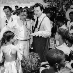 Welles gesticulando enquanto fala em área externa, rodeado por crianças