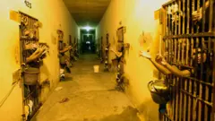 Corredor de presídio no Acre, com portas dos dois lados e braços para fora das celas