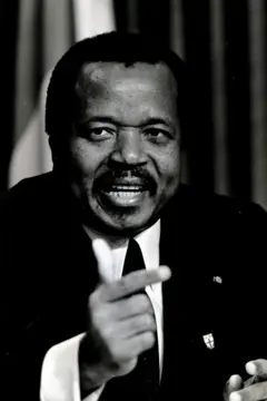 Fotografia em preto e branco do presidente dos Camarões, Paul Biya, com aparência mais jovem, vestindo um terno e fazendo um gesto com a mão direita