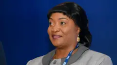 Jewel Cianeh Taylor wears a blue lanyard and grey jacket, gold earring at an economic forum in Poland, 2021. She has shiny black hair.