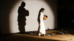 Latifatou Ouédraogo verse de l'eau d'une cruche dans des tonneaux pour l'ouverture de la première du premier acte de Là-bas ou Ici à l'Institut Français de Ouagadougou le 23 avril 2021.