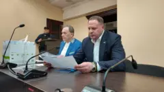 У приміщенні з бежевими стінами за столом сидять двоє людей. Обоє в піджаках і світлих сорочках, перед ними аркуші паперу, які вони тримають у руках і читають. На столі — мікрофони, папки та документи. За їхніми спинами стоїть озброєна людина в темній формі біля дверей.