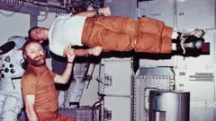 Jerry Carr demonstrates weightlessness by pretending to hold up Ed Gibson with one finger
