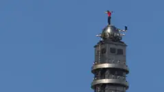 Алекс Хоннолд в красной рубашке стоит, раскинув руки, на шарообразном куполе, венчающем здание Taipei 101 