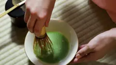 畳の上に茶碗を置き、茶筅(ちゃせん)で薄茶を点てている人の手元。茶碗の横には、漆塗りの黒い棗(なつめ、抹茶を入れる容器)の上に茶杓(ちゃしゃく)が置かれている