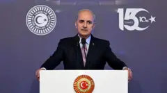  Numan Kurtulmuş ellerini kürsünün iki yanına koymuş konuşma yapıyor. Önünde TBMM forsu amblemi var. Kürsü beyaz.