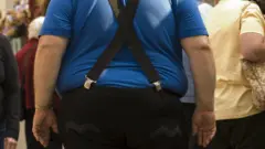 Homem obeso de costas, rodeado de outras pessoas