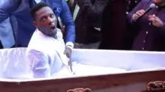 Pastor Alph Lukau stands next to the coffin and places his hand on the stomach of the man he is claiming to resurrect.