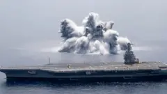 A large wall of water raising above the sea surface next to an US aircraft carrier due to the impact of explosion