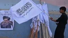 A vendor holds a Taliban flag next to a poster of Taliban leader Abdul Ghani Baradar as he waits for customers along a street in Kabul on August 27, 2021.