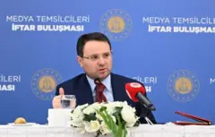 Akın Gürlek ardında medya temsilcileri iftar buluşması yazılı mavi bir panonun önünde oturuyor. Önünde çiçek ve mikrofon var. 