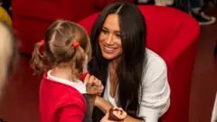 The Duchess of Sussex crouches receives a cake from a little girl at Windsor's Broom Farm Community Centre