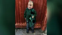Edward en uniforme escolar verde, con un maletín y está parado frente a una valla de madera