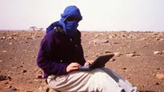 Jeff Greenwald em um terreno aberto e pedregoso, com rosto coberto por pano e laptop no colo