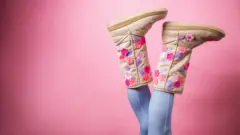 A pair of legs upside down, wearing skinny jeans and a pair of Ugg boots decorated with flowers - bright pink background.