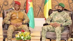 Burkina Faso president on di left wearing military uniform and red beret, Mali president sit for di right hand side wearing military uniform and green beret. Flags dey dia back
