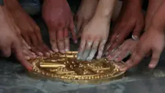 Several pairs of hands press down on a gold plaque that has been lain in the ground