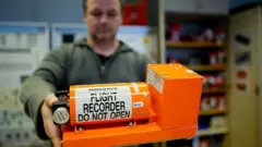 bir görevli elindeki parlak turuncu uçuş kayıt cihazını gösteriyor.