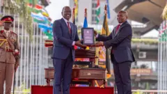 President Ruto dey hand ova a plaque to di new deputy as both of dem smile and one officer dey salute dem for back