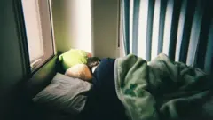 Pessoa branca de cabelos pretos de costas embaixo de um cobertor em uma cama