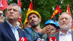 Saroı kasklı bir madenci konuşuyor, önünde mikrofonlar var. Yanında Özgür Özel duruyur