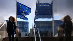 Pedestre desce escadarias em Bruxelas tendo como pano de fundo bandeiras da União Europeia hasteadas
